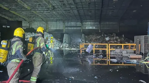 Mike Sudds The inside of a waste recycling centre where a fire has happened. Two firefighters are in the area of the building where the photograph has been taken and are holding a hose. There is a pile of rubbish and there looks to be smoke damage inside the building.