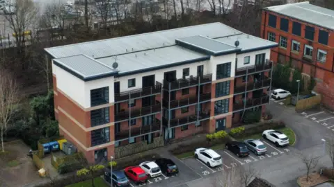 An aerial view of a multi-storey housing complex, featuring a flat roof, four storeys, and a number of cars parked in a car park running along the front of a building. Trees can be seen behind the building. 