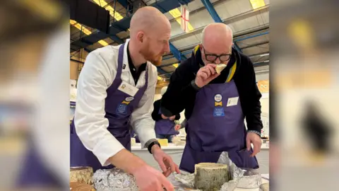 PA Media Two men wearing purple aprons, analysing some cheese at the award show. There is a low table in front of them full of different types of cheese. The man on the right is bald, wearing black thick framed glasses and is smelling a small chunk of pale cheese. The man on the left, bald with a red beard, is watching him intently. They appear to be in a large warehouse type building with high ceilings.