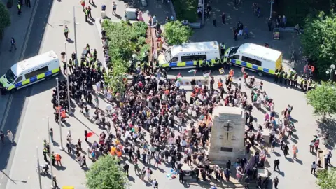 Avon and Somerset Police An aerial view of a large crowd of people gathering close to a war memorial. They are partially surrounded by three police vans and groups of police officers. The spaces is dotted with trees.