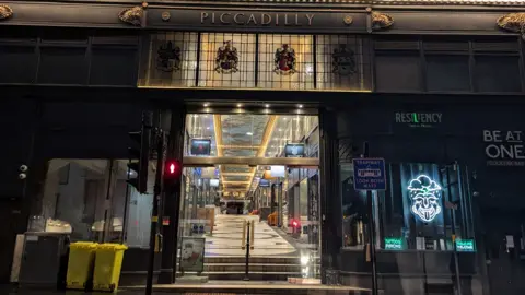 The entrance to Piccadilly Arcade in Birmingham - Piccadilly is written above the entrance in gold letters with automatic doors leading into a shopping arcade underneath.