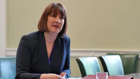 Reuters Rachel Reeves sits at a desk with empty chairs beside her. She's leaning forward and appears to be listening and is holding a piece of A4 paper