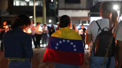 Relatives of political prisoners hold candles and posters during a vigil in Zamora, Venezuela, 26 February 2026. A woman, with her back to the camera, has a Venezuelan flag wrapped around her shoulders. Others hold candles.