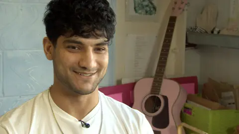 Ibrahim is wearing a cream t-shirt and is smiling at the camera. Behind him is a classical guitar leaning against some wooden boxes. 