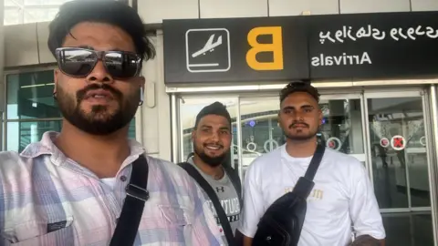 Three Indian men with sunglasses and shoulder bags standing in front of an airport in Tehran