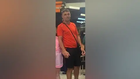 GOFUNDME A man is wearing an orange shirt and black shorts with the strap of a black bag horizontally across his body