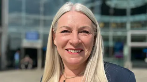 Jo Thewlis/BBC Kay Mason-Billig with grey, very straight, long hair, groomed eye-brows and pink tinted lips.