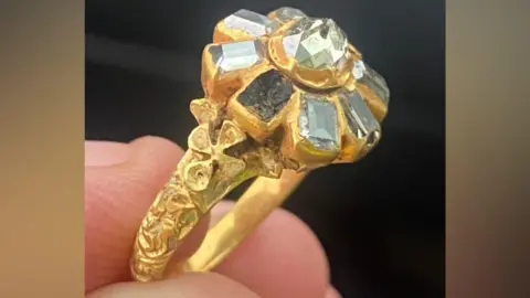 Stuart Hall A close up of a ring, which a man is holding between his fingers and thumb. The gold ring has eight diamonds which are arranged in a floral shape, although some are missing. The band of the ring is engraved.
