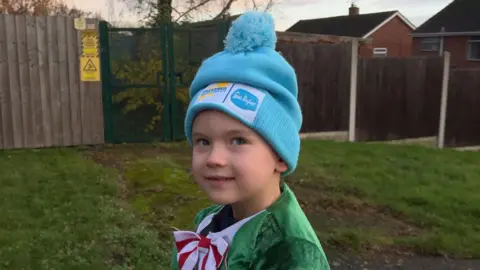 William looks up at the camera with a smile. He is wearing a green, red and white elf costume and an oversized baby blue bobble hat. 