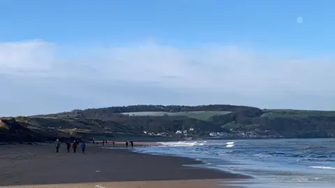 A wide sandy beach with small waves along the shoreline. People can be seen walking in the distance along with green hills and scattered buildings.