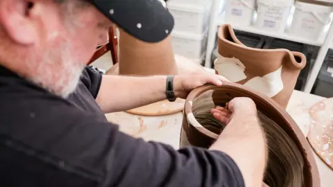 British Ceramics Biennial Johnny Vegas looks down at a piece of clay he is trying to shape. It is placed on top of a table with other pottery items.