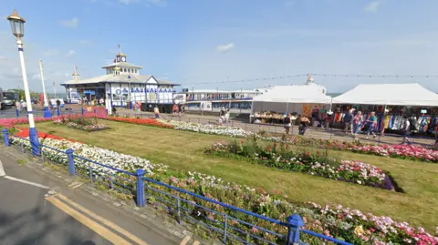 Google This screenshot, taken from Google Maps, shows Eastbourne Carpet Gardens in the late summer of 2024. The beds are populated with red, white and pink flowers and the lawn appears freshly cut and slightly scorched by the sun. The sky is clear blue and Eastbourne Pier is visible in the background.