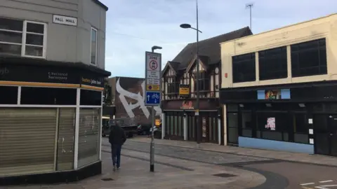 A shopping street that has several empty buildings. A man is walking away from the shot and there is a "to let" sign on one of the empty buildings on the right. The sky is a cloud but still pale blue. The road sign reads "Pall Mall". 