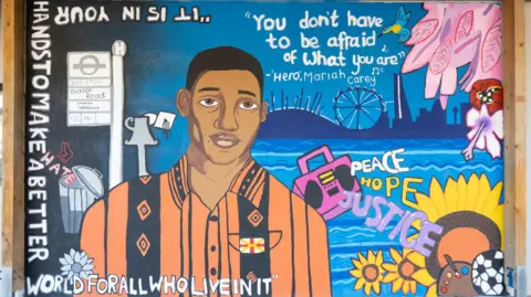 Eltham Hill School Mural showing Stephen Lawrence in an orange and black Jamaican style shirt with the words "It is in your hands to make a better world for all who live in it" written around the sides of the painting