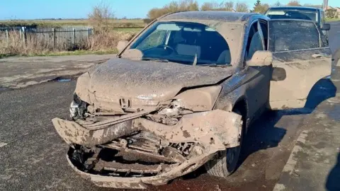 A mud splattered vehicle which has been damaged and crushed at the front underneath the bonnet. The vehicle is parked by the side of a road with a field in the background.