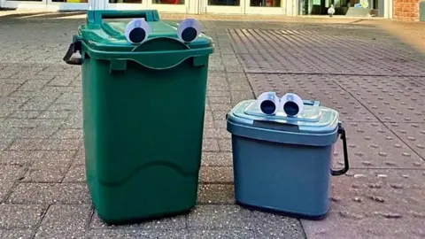 Two bins on concrete. One is large and green and the other one is smaller and blue. They both have googly eyes stuck on them 