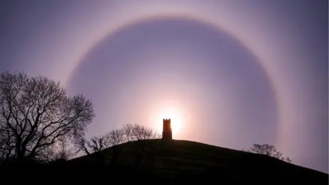 Josh Dury A halo of light is captured around the silhouette of Glastonbury Tor. The picture is taken from the bottom of the hill and the sun is rising in the distance. Silhouettes of trees and a hill are also visible.
