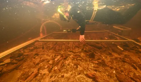 PA Media A diver floating in shallow water, holding what appears to be a metal frame just over the water bed. The diver appears to be examining a wooden platform nestled in the rocky, sandy bed.