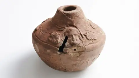 Small Roman pottery vessel with foliage and face. The vessel is a light terracotta colour and the vine leaves and face design is around the top curved section of the vase and there's a crack just by the face.