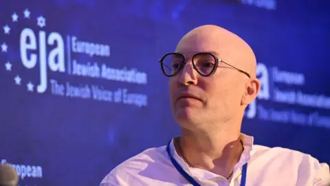 Alex Hearn Alex Hearn wearing glasses, a white shirt and a lanyard, looks to the left of the camera lens. Behind him is an advert for the European Jewish Association.  