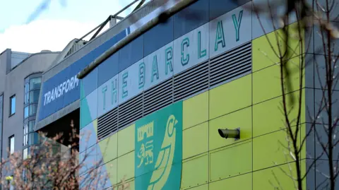 PA Media Carrow Road football stadium in Norwich with the yellow and green logo on thr side of the building.