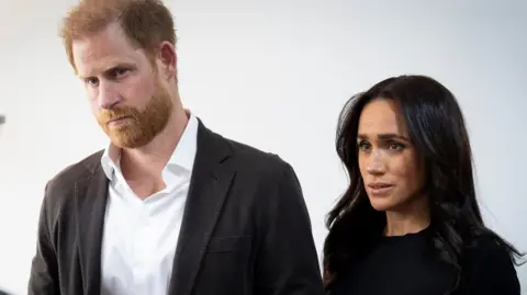 Harry and Meghan pictured in front of a white wall. Harry has short red hair and a beard. He wears a white collared shirt and a black open blazer. Meghan has medium-length black hair and wears a black long-sleeved top. 