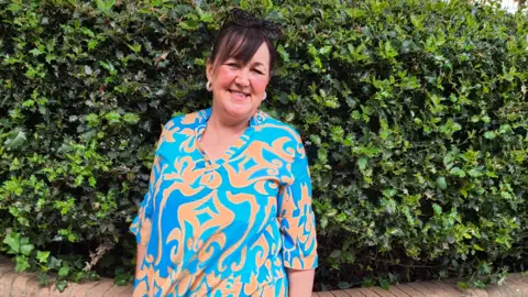 Louise Allen smiling at the camera. She is wearing a bright blue and orange blouse, she has sunglasses resting on the top of her head and is wearing white hooped earrings. She has dark hair with a fringe.
