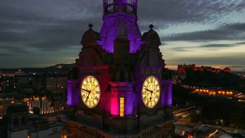 Guven Ozdemir/Getty Images A close up of the Balmoral Hotel clock showing two of its faces. It is lit up in a purple light and looks atmospheric because it is against a dark nights sky. In the background are the lights of the city's streets and Edinburgh Castle.