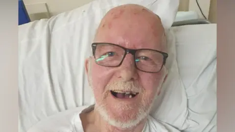 A man with white hair and glasses in a hospital gown lying in a hospital bed. 