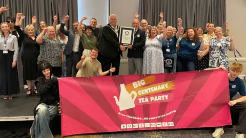 A group of people are stood with the arms raised. At the front is a pink banner which has white lettering that reads Big Centenary Tea Party.