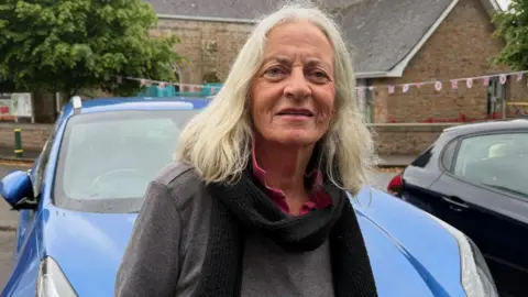 Mary Tunney is wearing a black scarf, pink t-shirt and grey jumper. She is pictured in St John village.