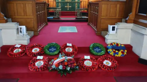 Crown Copyright The inside of a church with red velvet flooring and wooden choir stalls on either side. A series of poppy wreaths are laid on the steps. 