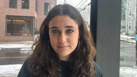 BBC Mia Tretta sits in front of a window in her dorm in Brown University with a street in the background with white snow on the ground. She is wearing a jumper and gold hoop earrings. 