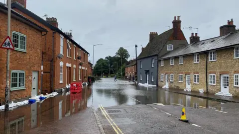 Louise Potter A road lined by cottage style houses with part of the road flooded, and houses lined by sand bags.