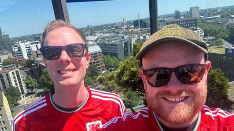 Photo of Matt Jones and Aaron Hill. Matt is on the left wearing a red Wales football shirt and has sunglasses on. Aaron is on the right, and is wearing a red Wales football shirt and sunglasses as well as a khaki hat