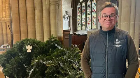 A man wearing a Gillet and green long sleeve top stands next to a large Christmas tree, which has fallen over, inside a church.