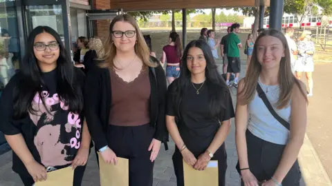 Phil Harrison/BBC Four female students stand outdoors in a school playground holding large brown envelopes on a sunny day.