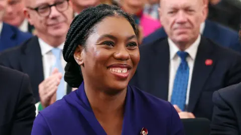 EPA-EFE/REX/Shutterstock Kemi Badenoch smiling. She wears a purple dress with a red poppy pin, while a crowd is sat behind her, with two men's faces visible.