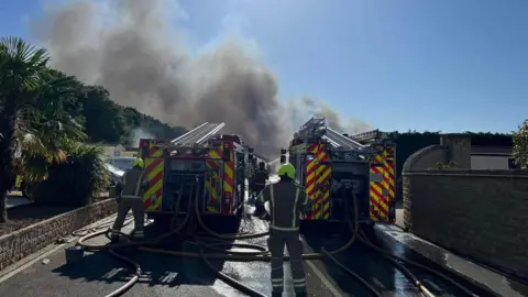Oxfordshire Fire and Rescue Service Two fire engines in the foreground parked on a road with firefighters working at the rear of the vehicles. In the background smoke can be seen pouring into the sky.