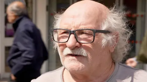 Robert Duggan is an older man with white hair, a white moustache and wearing black varifocal glasses.