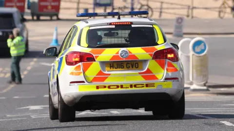 A police car with its hazard lights flashing is parked on a road that leads to a beachfront. There is a man wearing a high-viz jacket blurred in the background and a mixture of bollards and a blue traffic cone. 