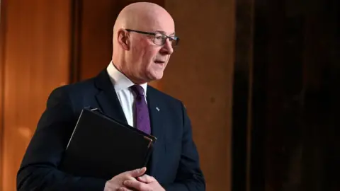 John Swinney, who is bald and wearing glasses, walks while holding a black folder under his arm. He is wearing a dark suit, white shirt and purple tie. 