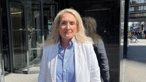 BBC A woman with blonde hair and glasses, in a blue and white striped shirt and white jacket, stands by a black marble column with glass rotating doors in the background