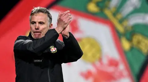 Phil Parkinson applauds the fans with a Wrexham flag in the background
