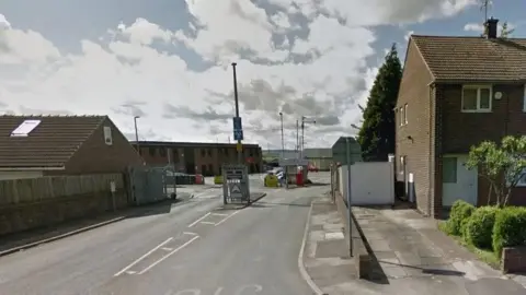 Street view of an entrance to a depot on a suburban road. A two-lane road leads toward security gates and barriers in the center of the image. Brick residential houses line both sides of the road, with a fenced boundary on the left and a pavement on the right. Several signposts, security equipment, and parked vehicles are visible beyond the gate. The sky is partly cloudy with bright sunlight casting shadows across the road and buildings.