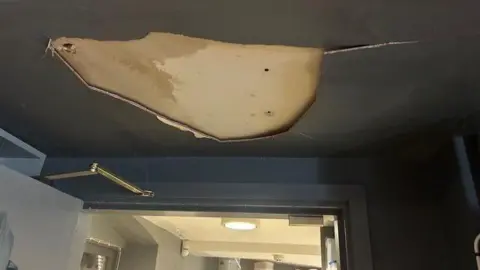 Stroud Valleys Artspace A large hole in the dark paint and plaster on a ceiling, revealing sodden floorboards above after a pipe burst. The doorframe, wall and ceiling are all painted with dark grey paint, and a door can be seen leading to a lighter space.