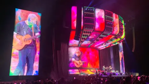 Richard Haugh/BBC Ed Sheeran performs on a large stage in Chantry Park in Ipswich. He can be seen projected on to multiple screens around the stage. He can be seen in the distance performing on the stage.