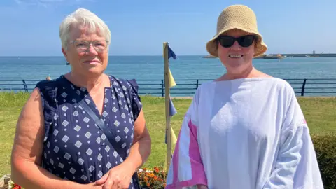 Kathryn Strain (left) and Bridie McElhill. Kathryn has short white hair and a blue dress while Bridie has a straw hat and is wearing a long bathing robe. They are standing outside on a sunny day, behind them is grass and further in the distance the sea.