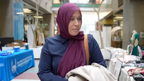 Eloise Alanna/BBC An image showing Lisa Robicheau wearing a purple headscarf. She is looking down towards the left with a blank expression. She is wearing a blue collared long-sleeve shirt, with a bag strap visible on her left shoulder. She is walking through a university audtorium, and she is surrounded by tables with various displays.