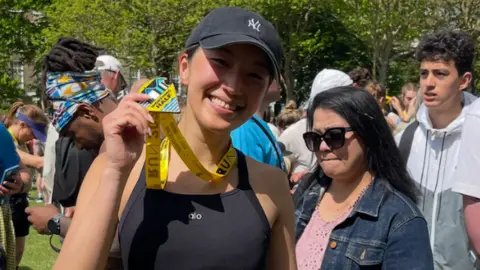 Andy Allen A woman with a black vest and baseball cap, with a sheet of numbers attached to her shorts, holding a runner's medal. 